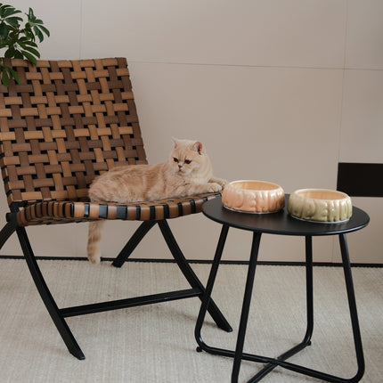 Cat lying on a woven chair next to a small round table with decorative items.