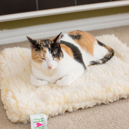 Calico cat lying on a fluffy white pet mat with a visible brand tag.