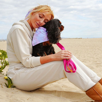 Woman sitting on a sandy beach holding a small dog with a pink leash.