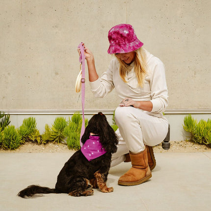 Woman in white outfit and pink hat with a dog wearing a pink bandana on a concrete surface.
