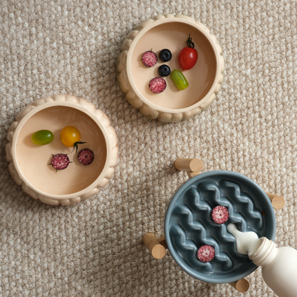 Two wooden bowls with small toys and a blue textured bowl being filled with water from a bottle on a beige surface.
