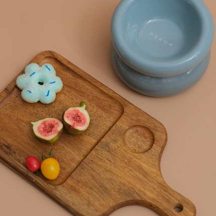 Wooden cutting board with sliced figs, a blue flower-shaped object, and a small stack of blue bowls on a beige background.