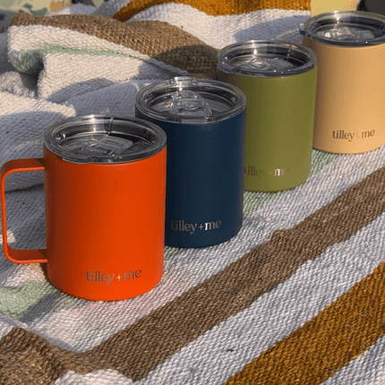 Four insulated mugs in orange, navy, green, and beige on a textured fabric surface with 'tilley + me' branding.